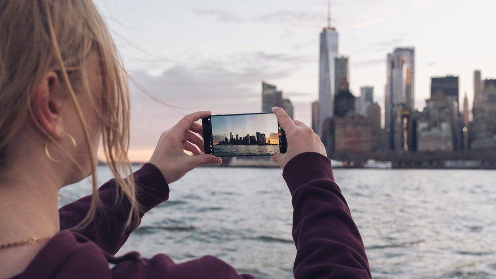 Image of a woman taking a photo of the Manhattan skyline at dusk with her phone