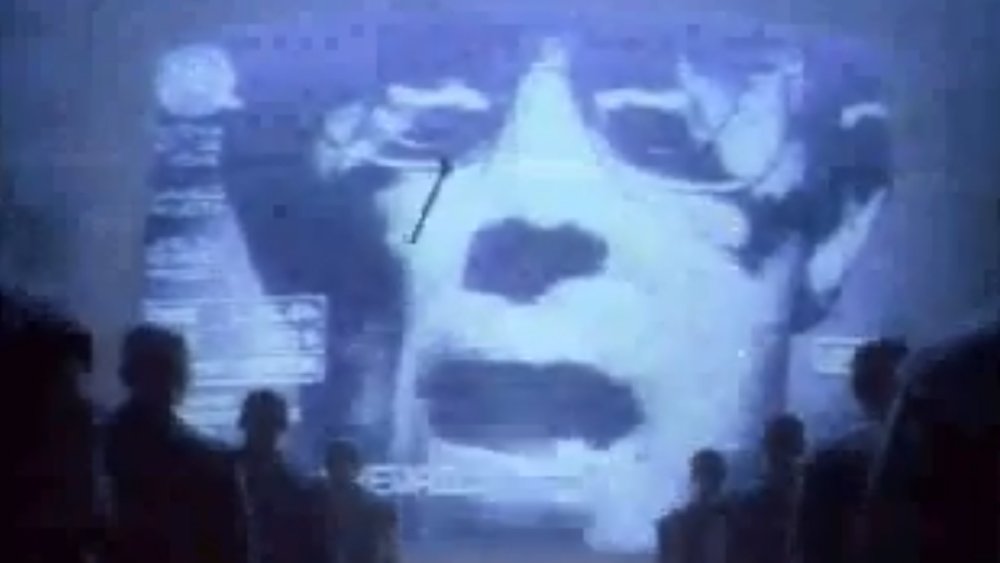A cowed audience are lectured by a face on a giant screen